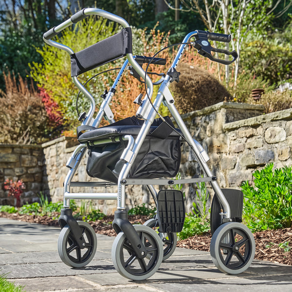 NRS 2 in 1 rollator and transit chair with padded seat and storage, on a garden path by stone walls and plants, angled side view.