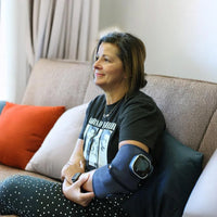 Woman sitting on a couch with an arm brace, Lifemax 3 in 1 Heated Vibration Massager beside her, front view.
