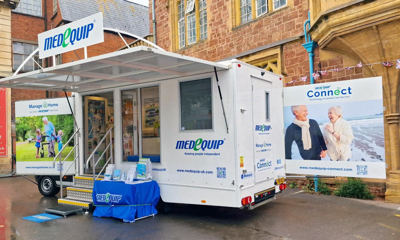 Medequip mobile unit parked on a street outside a historic building, with promotional signs for Manage At Home and Medequip Connect. The unit features branded banners and a display table with leaflets, welcoming visitors to learn more about independent living and care technology.