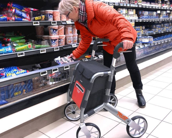 Trust Care Let’s Shop Rollator in black being used by an older adult to pick groceries from a fridge shelf, in use view.