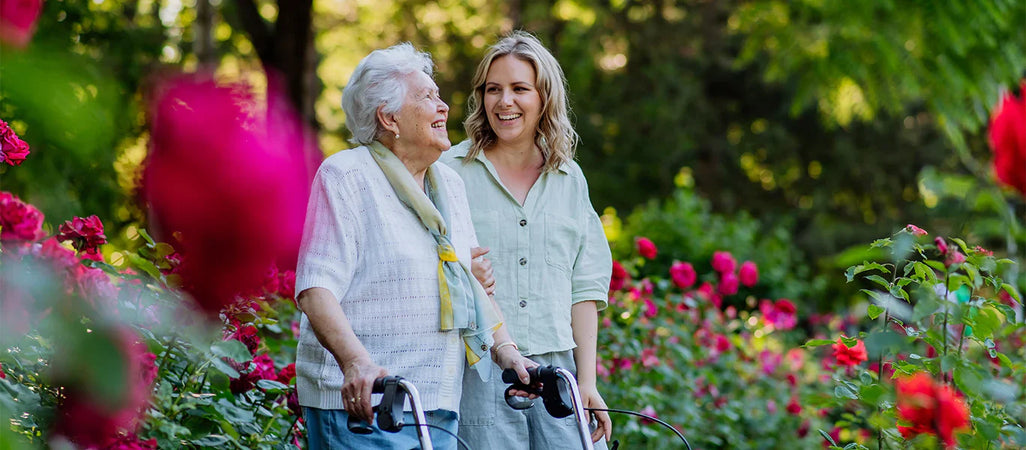 Elderly woman using a walker enjoys a stroll through a vibrant rose garden with a younger woman, demonstrating the product in an uplifting outdoor setting.