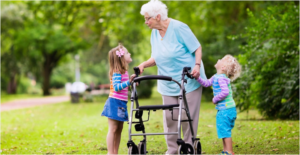 Elderly woman using a rollator walker in a park, smiling and holding hands with two young children, on a sunny day surrounded by green trees and grass.