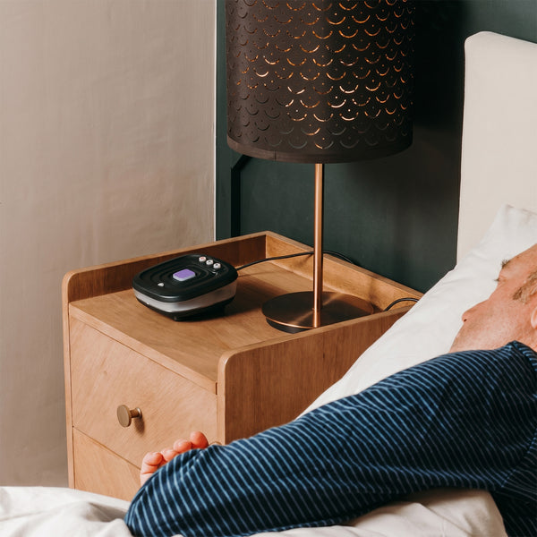 Older adult sleeping beside wooden nightstand with Ravencourt Bide Fall Prevention Device in use, bedside angle.