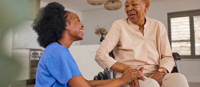 An elderly woman sits in a wheelchair, smiling and engaging with a younger woman who is demonstrating a product to her. They are holding hands and laughing in a bright, modern home setting, suggesting comfort and ease of use.