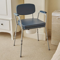 NRS Dovedale Comfort Commode with armrests and padded seat, placed in a carpeted room next to a cabinet and laundry hamper, side view.