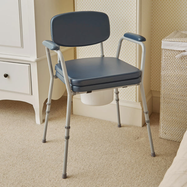 NRS Dovedale Comfort Commode with armrests and padded seat, placed in a carpeted room next to a cabinet and laundry hamper, side view.