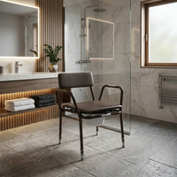 NRS Dovedale Extra Low Commode in a modern bathroom setting with marble walls, glass shower and wooden towel shelves, front view.