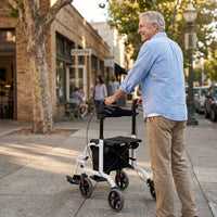 Older man walking with Aidapt Duo Deluxe Rollator on pavement near shops and trees, in use view.