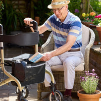 Man outdoors placing book in NRS Freestyle 4 Wheel Rollator basket next to potted plants, in use view.