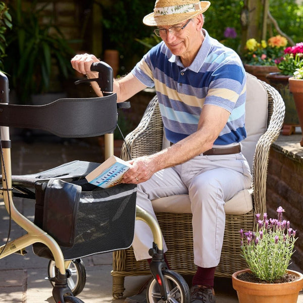 Man outdoors placing book in NRS Freestyle 4 Wheel Rollator basket next to potted plants, in use view.