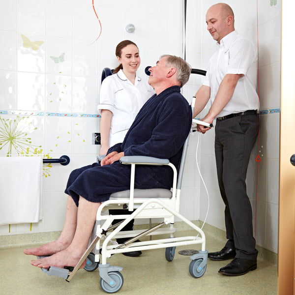 Prism Medical Freeway T80 Reclining Shower Chair in use with two caregivers assisting an older man in a tiled bathroom, side view.