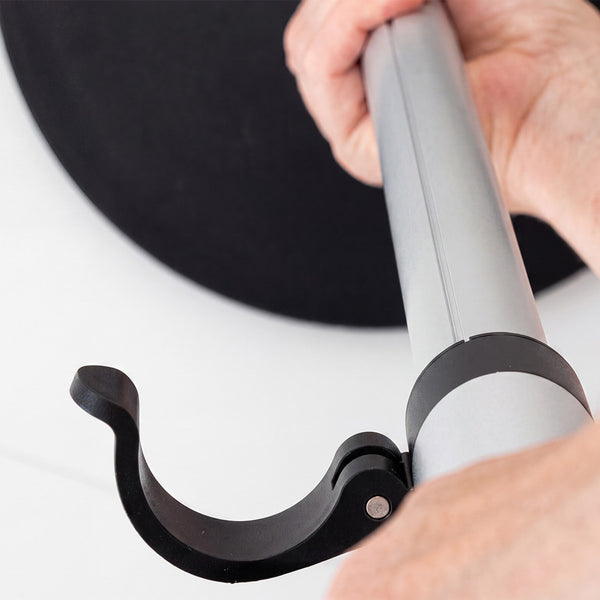 Hand holding a GRIPO Support Pole with black hook, near a round black object in the background, close-up.