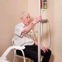 Elderly man on shower chair holding GRIPO Support Pole in bathroom with handheld showerhead, in use view.