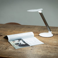 Halo Go 2 Magnifying Lamp by The Daylight Company next to an open magazine on a wooden table, plain background, front view.