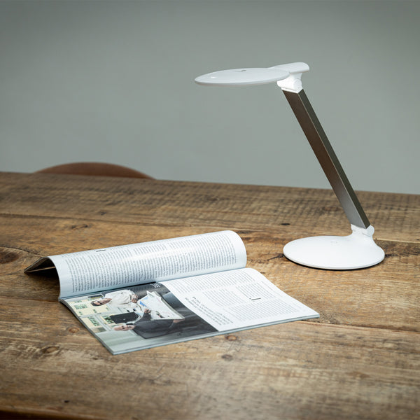 Halo Go 2 Magnifying Lamp by The Daylight Company next to an open magazine on a wooden table, plain background, front view.