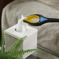 Helping Hand Handi-Grip Pro picking up a tissue from a box on a glass table beside a fern, in use view.