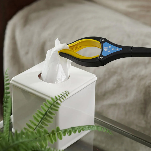 Helping Hand Handi-Grip Pro picking up a tissue from a box on a glass table beside a fern, in use view.