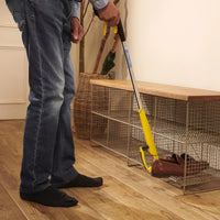 Helping Hand Shoe Helper used to pick up a brown shoe from under a wire bench on wooden floor, in use view.