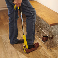 Person uses a Helping Hand Shoe Helper to put on brown shoes next to a metal shoe rack, in use view.