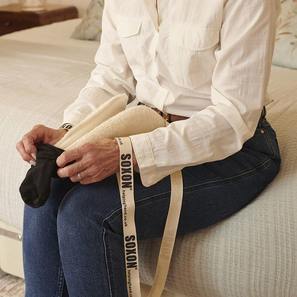 Person using a Helping Hand Soxon Sock / Stocking Aid to put on a sock while seated on a bed, in use view.