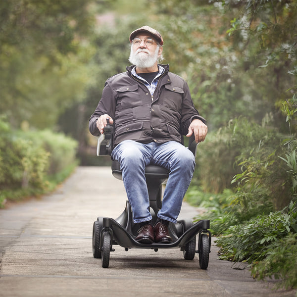 Older man using Urathon Hero Carbon Fibre Powerchair on a garden path with greenery and trees, in use view.