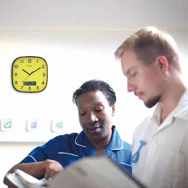 Lifemax High Contrast Day-Date Wall Clock showing 8:14 above two medical professionals reviewing a file in a bright room, in use view.