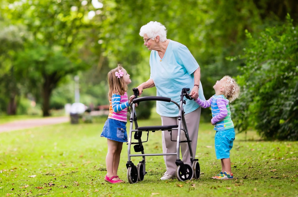 Elderly woman using a rollator walker in a park, smiling and holding hands with two young children, on a sunny day surrounded by green trees and grass.