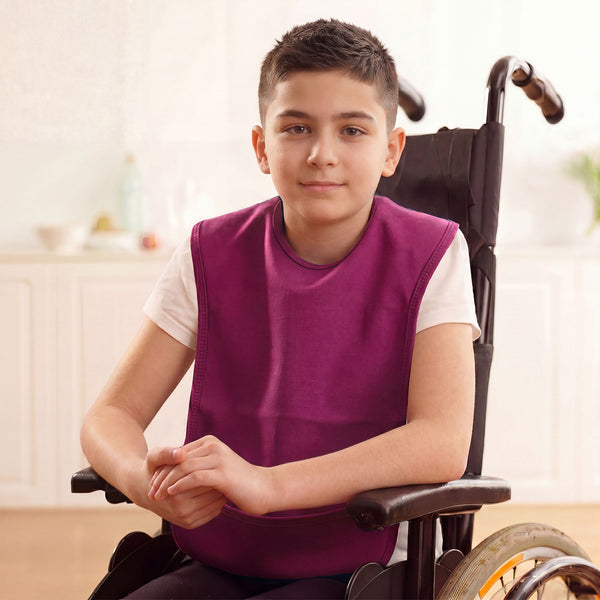 Boy in wheelchair wearing Care Designs Junior Tabard Style Bib, machine washable, sitting indoors, neutral expression, front view.