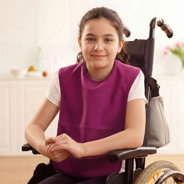 Young girl in wheelchair wearing Care Designs Junior Tabard Style Bib, indoors, smiling with soft background, front view.