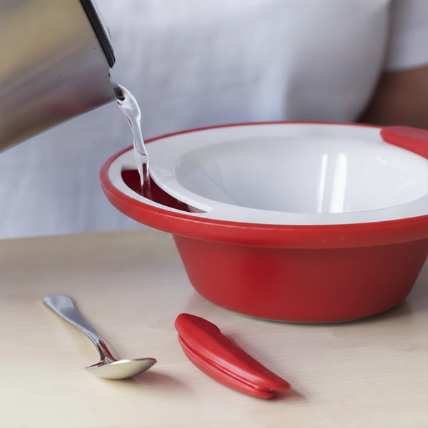 Person pouring liquid into an ORNAMIN Keep Warm Bowl with spoons and a red item on the table, in use view.