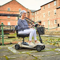 Older woman using the Motion Healthcare Lithilite Scooter with puncture-proof wheels outdoors by a pond and brick buildings, in use view.