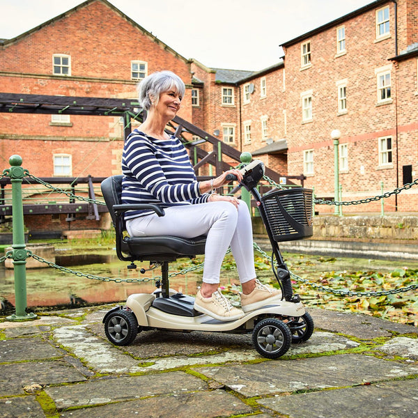 Older woman using the Motion Healthcare Lithilite Scooter with puncture-proof wheels outdoors by a pond and brick buildings, in use view.