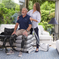 Mangar ELK Lifting Cushion in use to help a man with prosthetic legs, assisted by a woman, modern living room setting, side view.