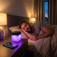 Lifemax Mood Light Bluetooth Speaker on a nightstand next to a glowing lamp and smartphone, man sleeping in bed, in use view.