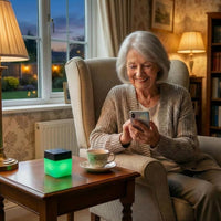 Lifemax Mood Light Bluetooth Speaker on table next to elderly woman in armchair with phone and teacup, in use view.
