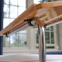 NRS Divan Overbed Table with height adjustable frame and chrome legs positioned beside windows, side view.