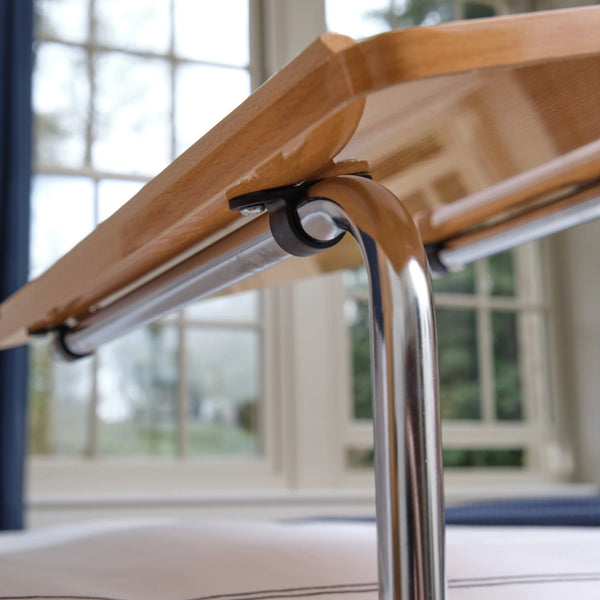 NRS Divan Overbed Table with height adjustable frame and chrome legs positioned beside windows, side view.