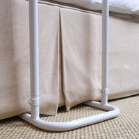 NRS Divan Overbed Table, height adjustable and wheelchair accessible, shown beside a bed with a fabric skirt on woven carpet, side view.