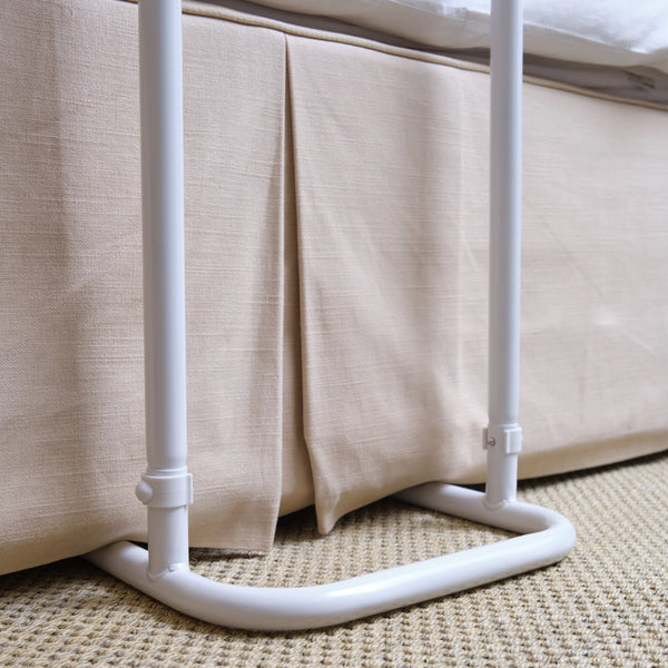 NRS Divan Overbed Table, height adjustable and wheelchair accessible, shown beside a bed with a fabric skirt on woven carpet, side view.