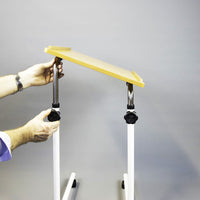 Person adjusting NRS Overbed/Overchair Table with white legs and wooden top, plain background, in use.