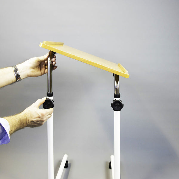 Person adjusting NRS Overbed/Overchair Table with white legs and wooden top, plain background, in use.