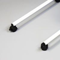 Pair of white metal rods with black caster wheels from NRS Overbed/Overchair Table on light grey surface, close-up.