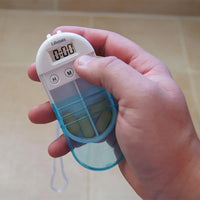 Hand holding Lifemax Pendant Pill Box with Reminder, pills visible inside and digital display showing 0:00, in use close-up.