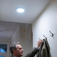 Lifemax Rechargeable Motion Ceiling Light installed above man hanging coat on wall hook in hallway, in use.