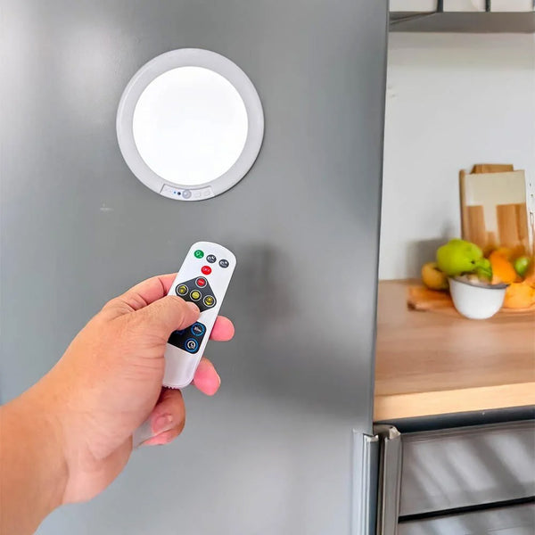 Hand holding remote pointed at Lifemax Rechargeable Motion Ceiling Light mounted on fridge, fruit and cutting board in background, in use view.