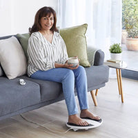Woman on sofa holding mug using Revitive Prohealth foot massager, product in use, side view.