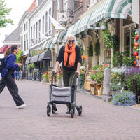 Older woman with white hair and orange scarf using a Rollz Air Carbon Fibre Rollator walking past flower-lined shops, in use view.