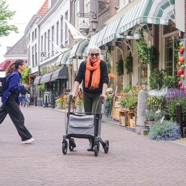 Older woman with white hair and orange scarf using a Rollz Air Carbon Fibre Rollator walking past flower-lined shops, in use view.