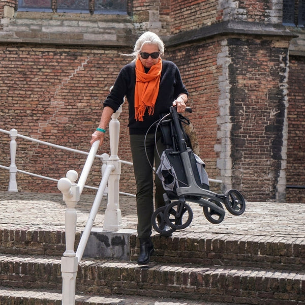 Older woman with grey hair and orange scarf carrying a Rollz Air Carbon Fibre Rollator down steps outside brick building, in use angle.