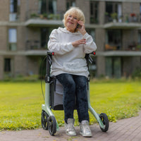 Elderly woman using a coal black carbon fibre Rollz Air rollator, talking on her mobile phone outside an apartment building, in use view.